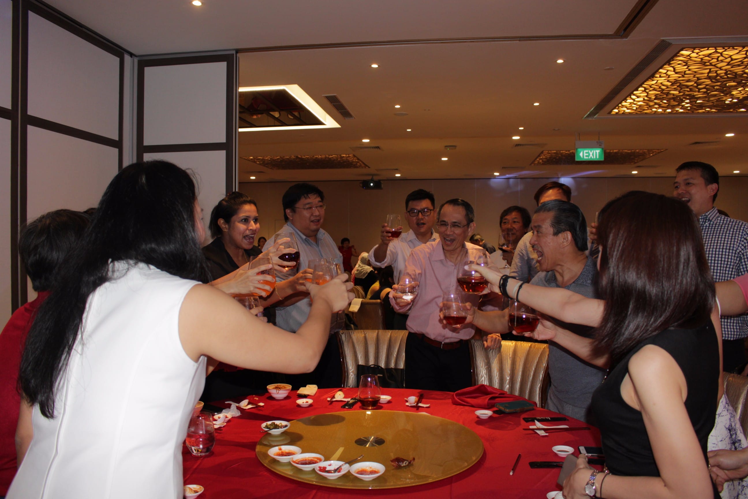 A group of people raising wine glasses in a toast around a red-clothed round dining table in a banquet hall.